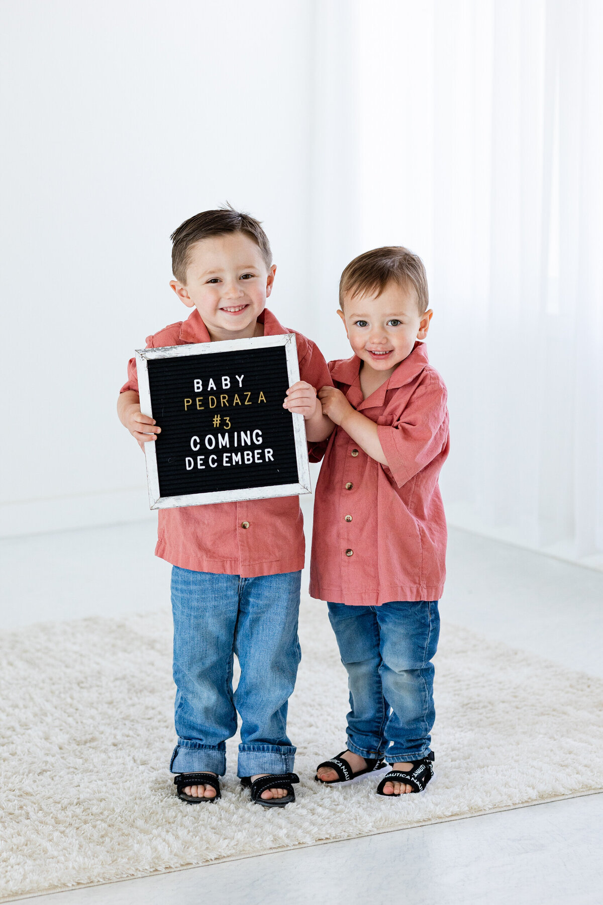 Two little boys smiling at the camera and holding a sign that reads, "Baby Pedraza #3 Coming December".
