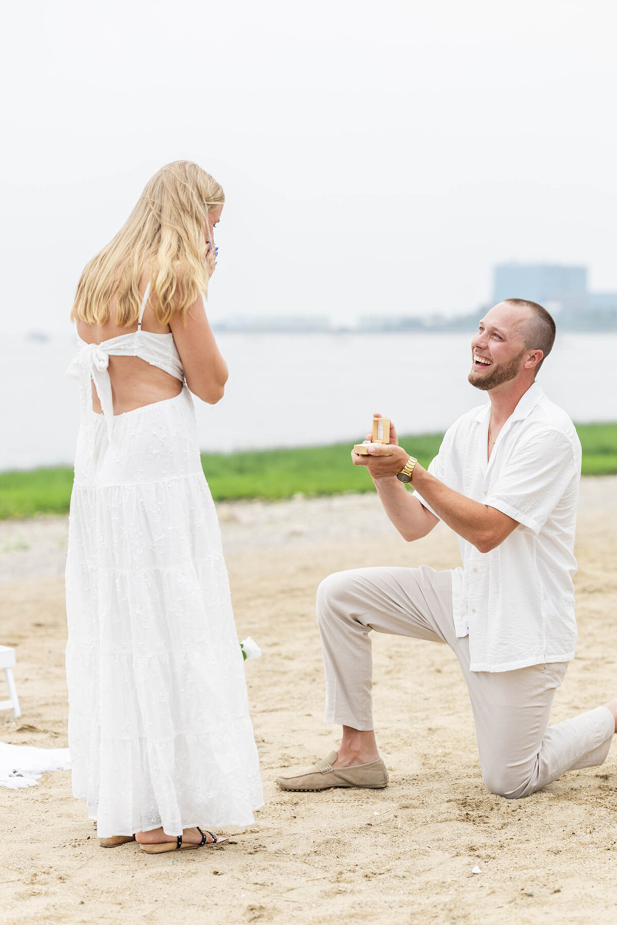 Woman wearing a white dress bends over covering her mouth as a man wearing a cream shirt and tan pants is kneeling on one knee in front of her holding an opened box with a ring displayed during a Coastal Connecticut engagement session. 
