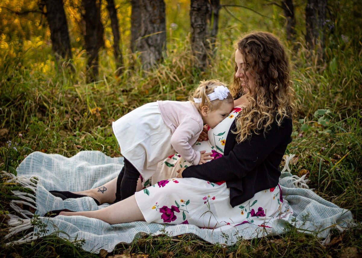 pregnant mom and her toddler daughter sit on a picnic blanket in a field in Edmonton
