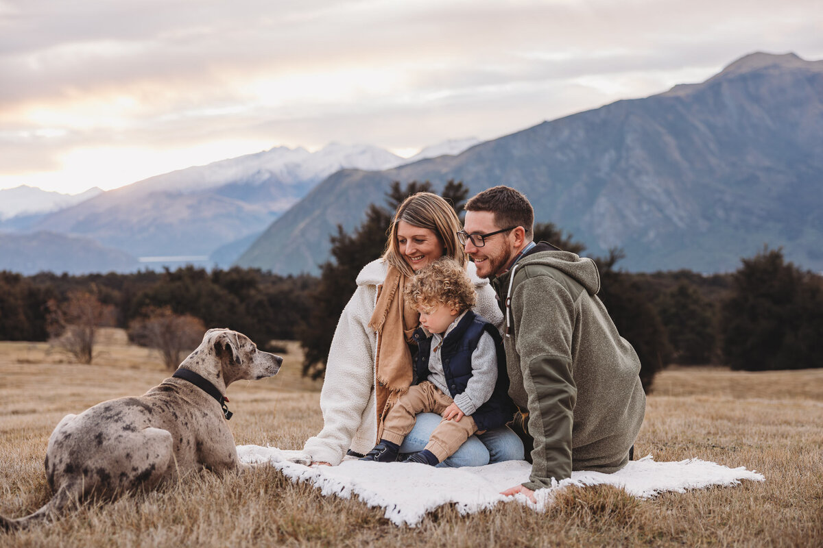a family sitting on a blanket looking at their dog laying in the grass next to them with the wanaka mountains in the background  during their family photoshoot in wanaka