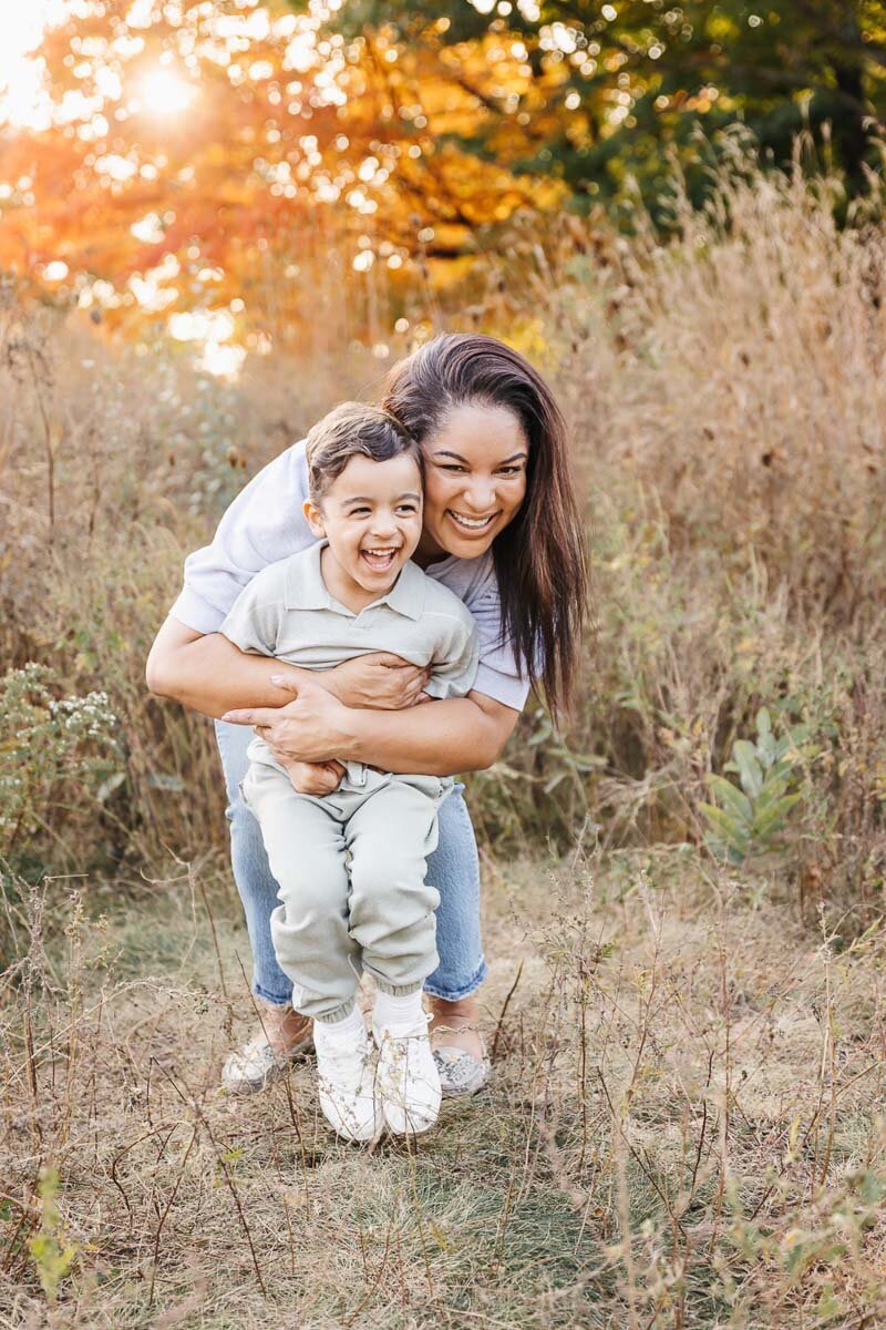 A woman smiles while hugging a young boy from behind amidst the tall grass and vibrant autumn foliage, capturing the essence of family photography in Toronto
