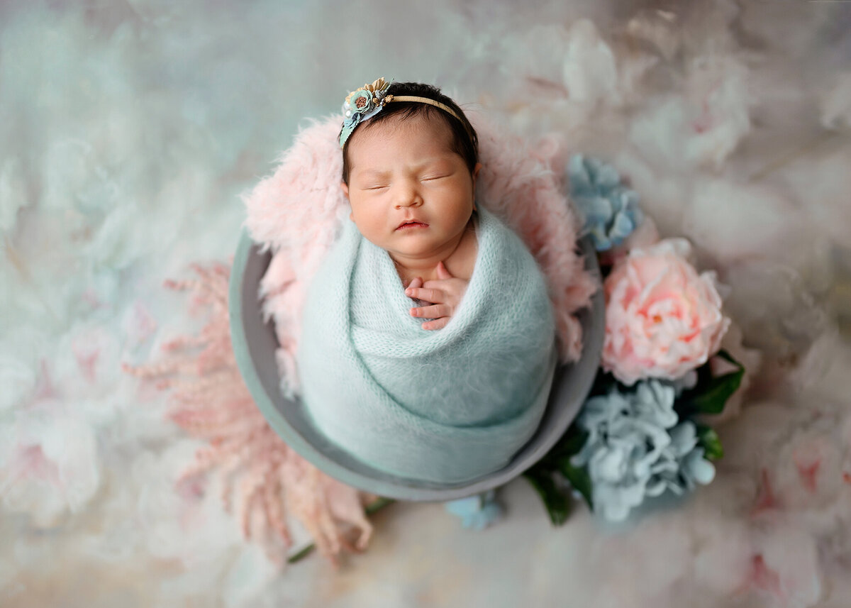 little baby girl in a soft blue swaddle sleeping in a gray bowl with pink and blue flowers surrounding her