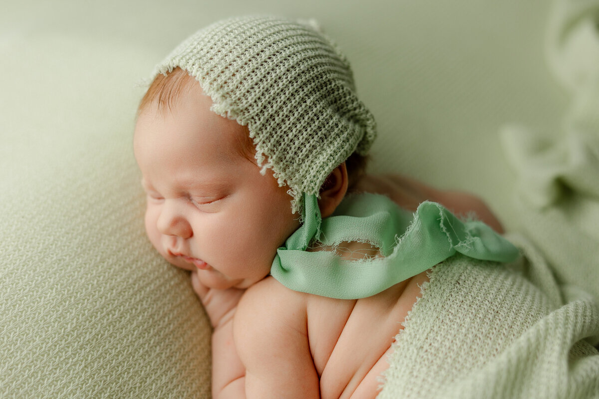 Close-up of a newborn baby resting on a sage green knit blanket, wearing a matching bonnet and soft wrap. Captured by Inland Empire newborn photographer Kaitlyn Dawn Photography.