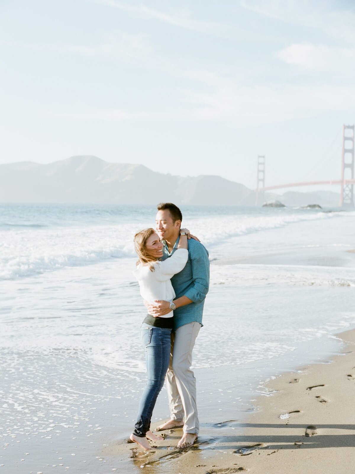 Tall handsome Asian man hugs his pretty fiancee at Baker Beach in front of the Golden Gate Bridge.