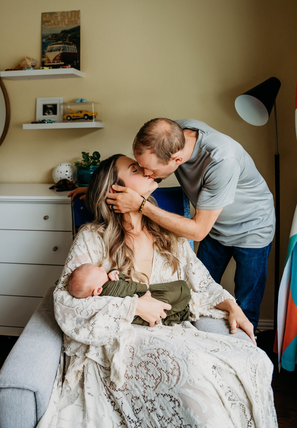 Newborn Photographer, woman sits and holds baby, husband leans in to kiss her