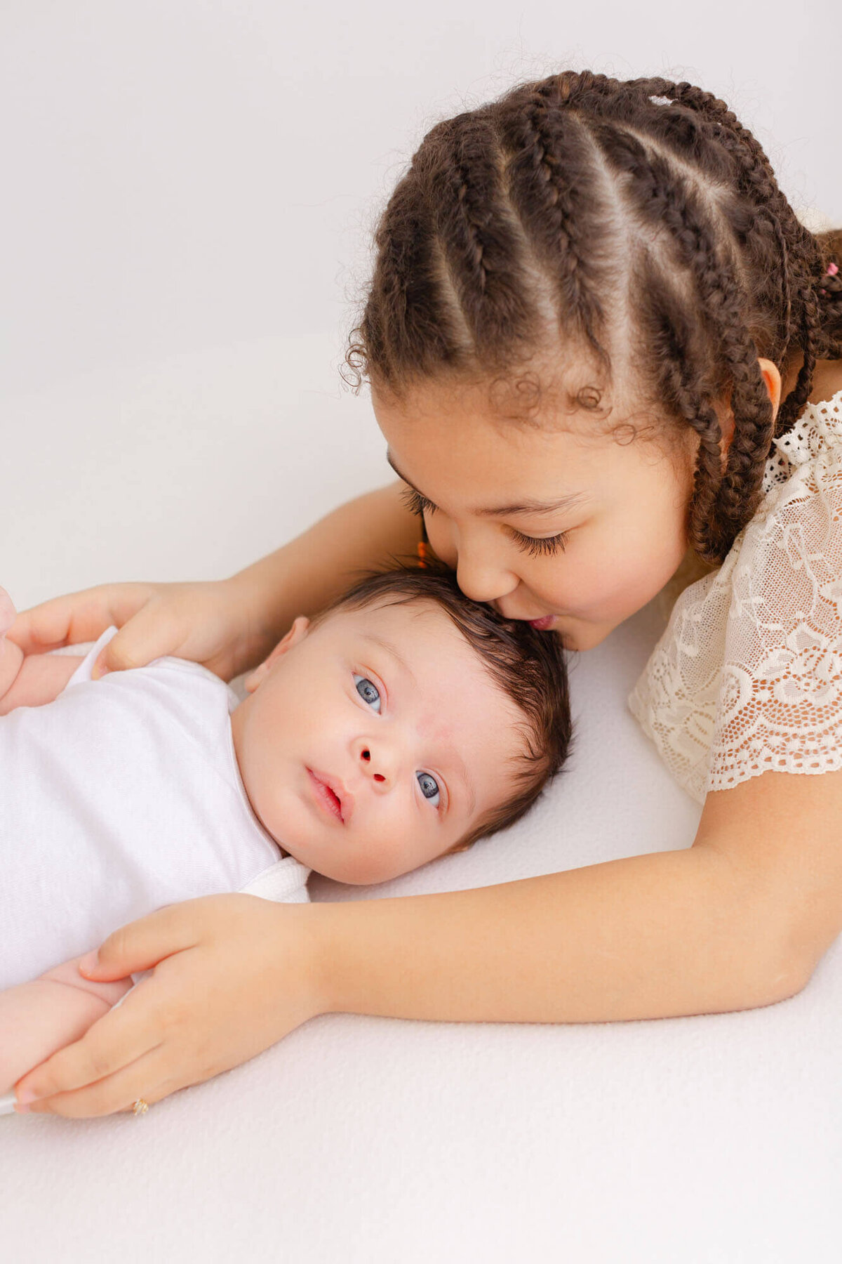 Baby looking at the camera with big blue eyes while sister kisses the top of his head from behind him and holds him in her arms.