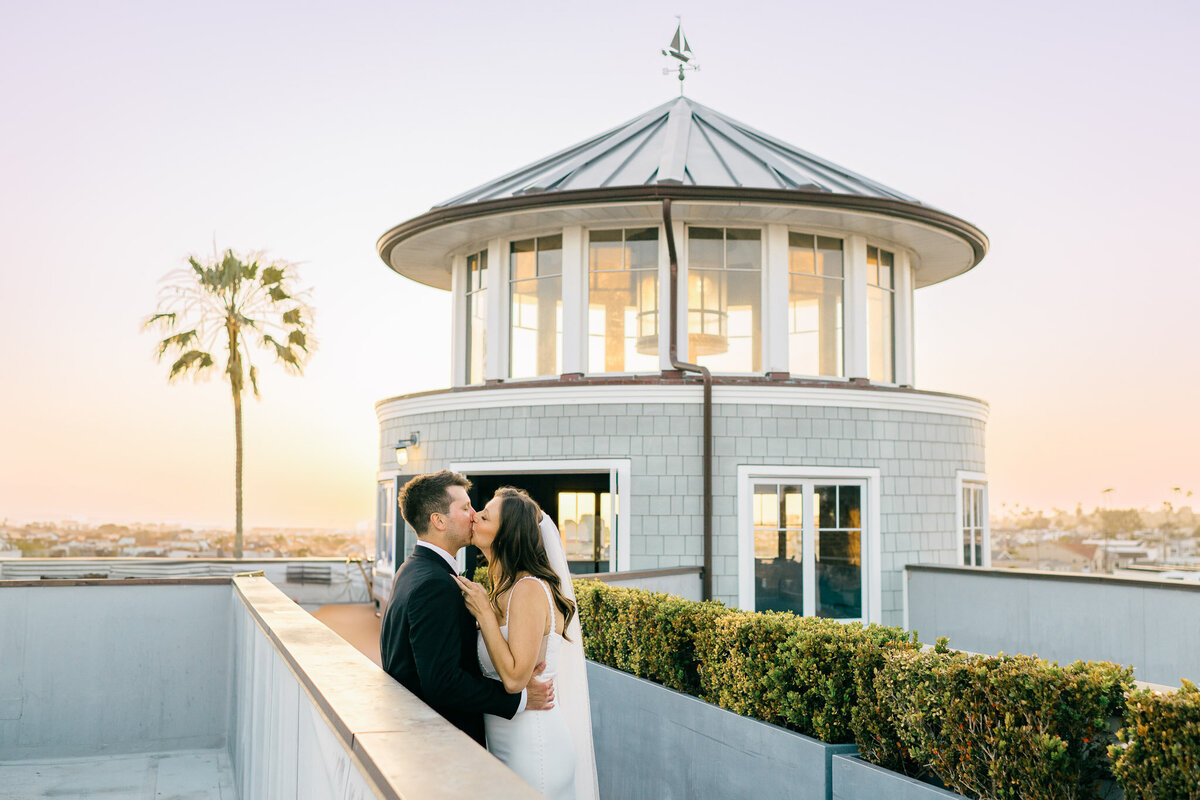 The bride and groom share a sunset kiss atop Lido House in Newport Beach, with a stunning panoramic ocean view behind them