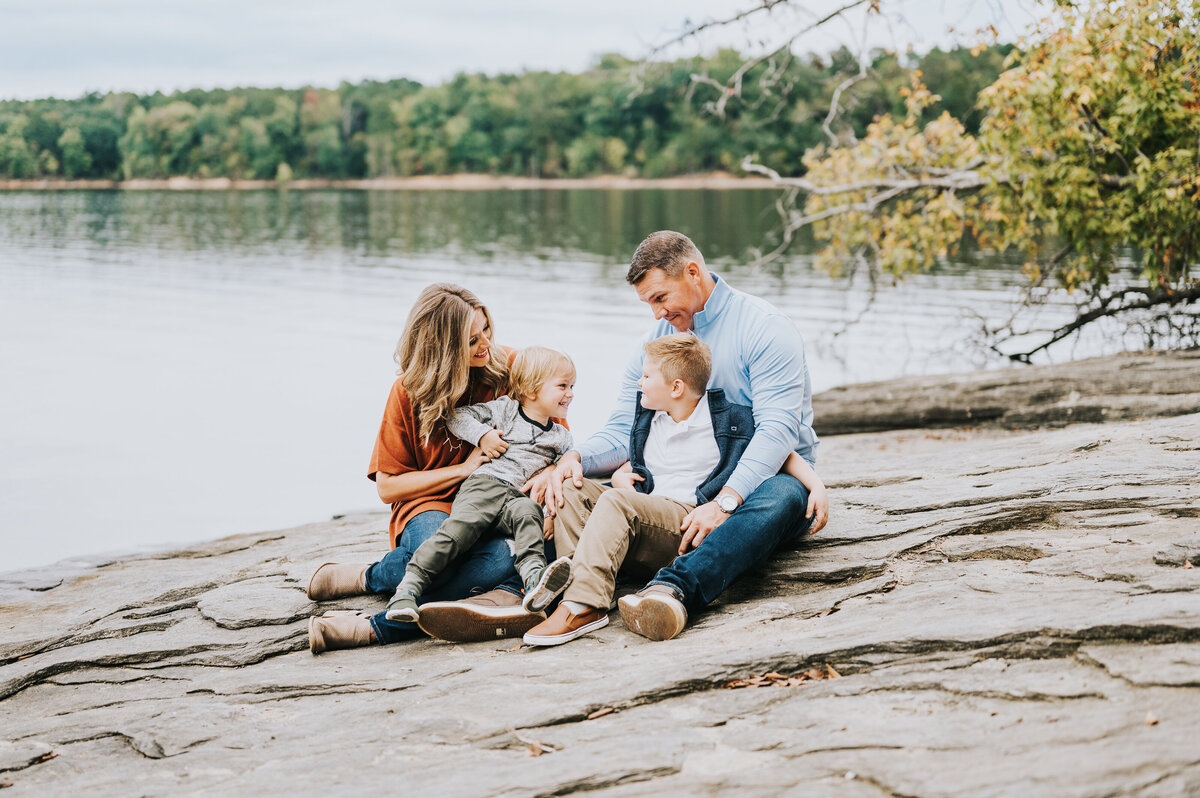 Young family with 2 boys laugh together beside a lake during family session