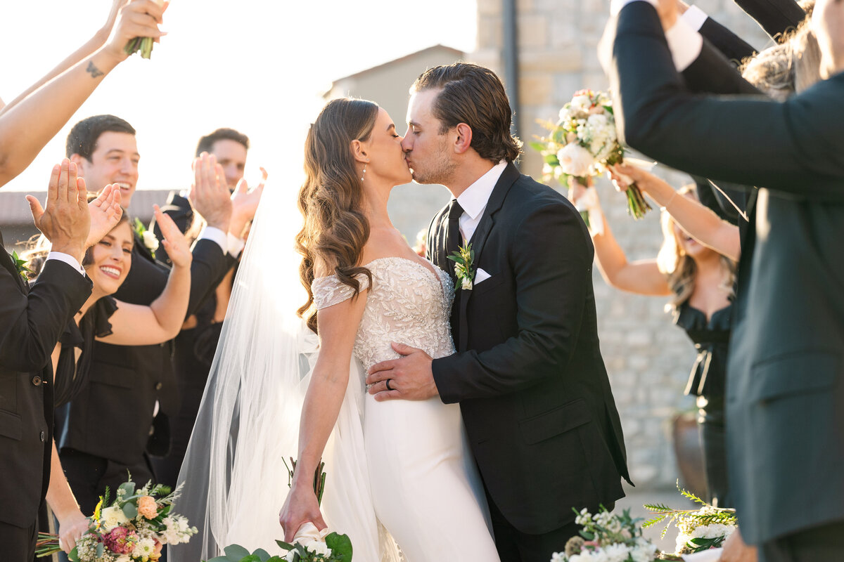 A joyful kiss between the bride and groom as their wedding party celebrates around them, captured during a golden hour celebration in Westlake, Texas.