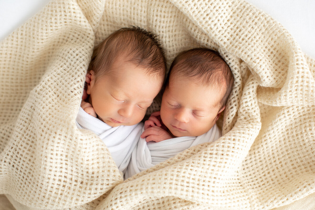 Newborn twins sleeping side by side in a woven basket, swaddled in cozy blankets and nestled close together.