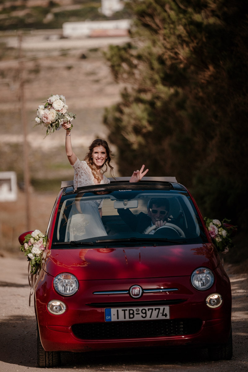 Photo de mariage à Paros en Grèce, avec un couple dans une voiture décapotable, la mariée agite son bouquet.
