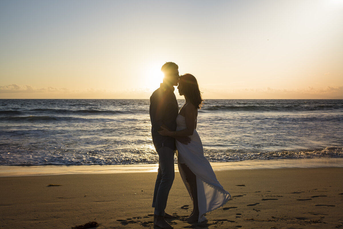 A silhouette of a bride and groom on the beach. The groom kisses his bride on the forehead.