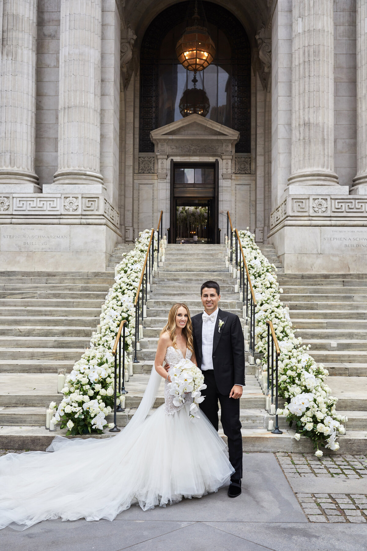 This enchanting moment was captured at the iconic New York City landmark, The New York Public Library, by the talented photographer Christian Oth, with videographers East West Films. In the photograph, a radiant bride and groom are captured standing on the steps of the grand building, surrounded by elegant white flowers. The bride looks stunning in her flowing white gown as she holds a delicate bouquet, while the groom exudes sophistication in his black suit and stylish boutonniere. The scene is beautifully framed by the majestic columns and intricate architecture of the library, adding a touch of grandeur to the couple's special day.