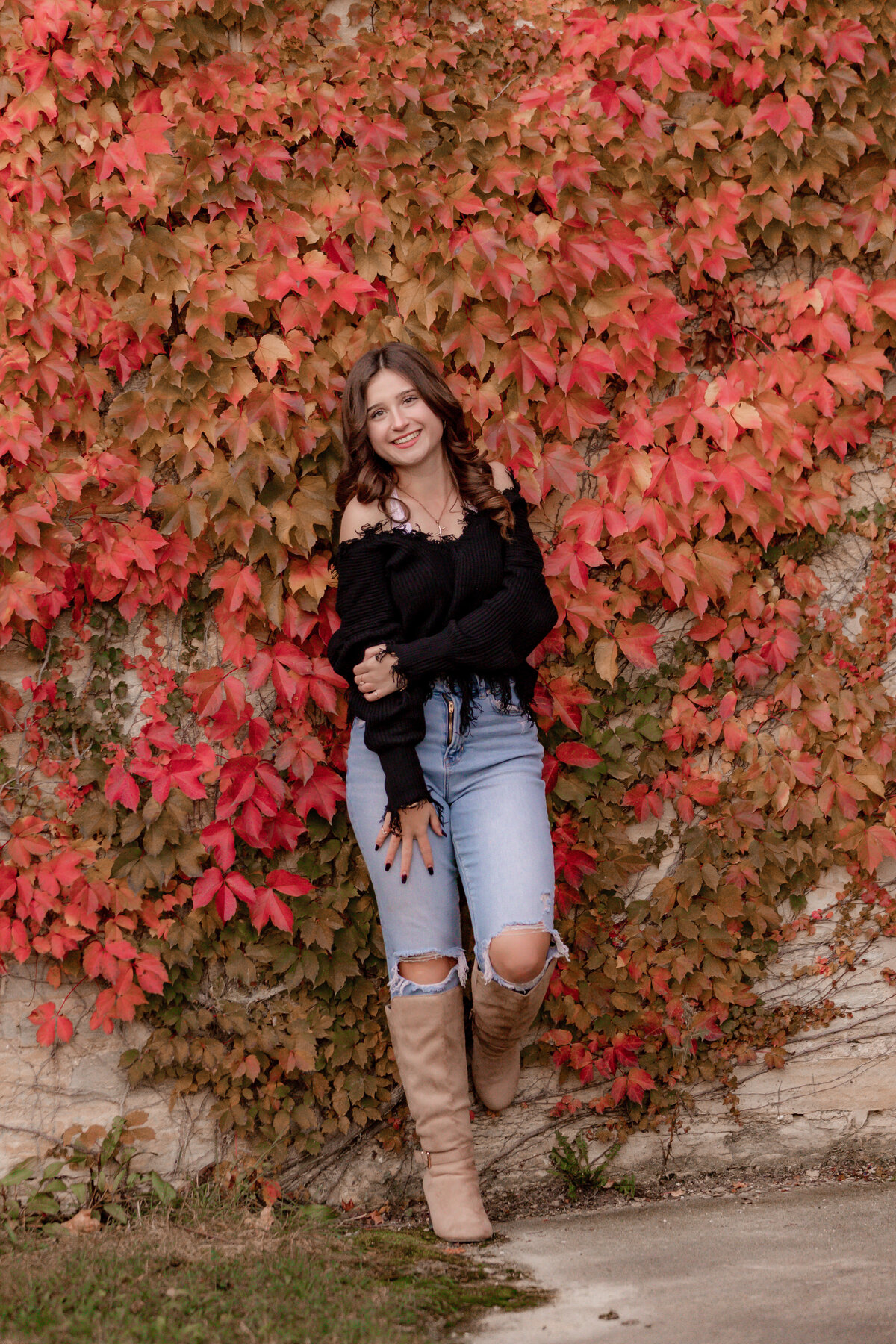A brunette leans against a fall colored ivy wall with one foot up and her hand across her body.