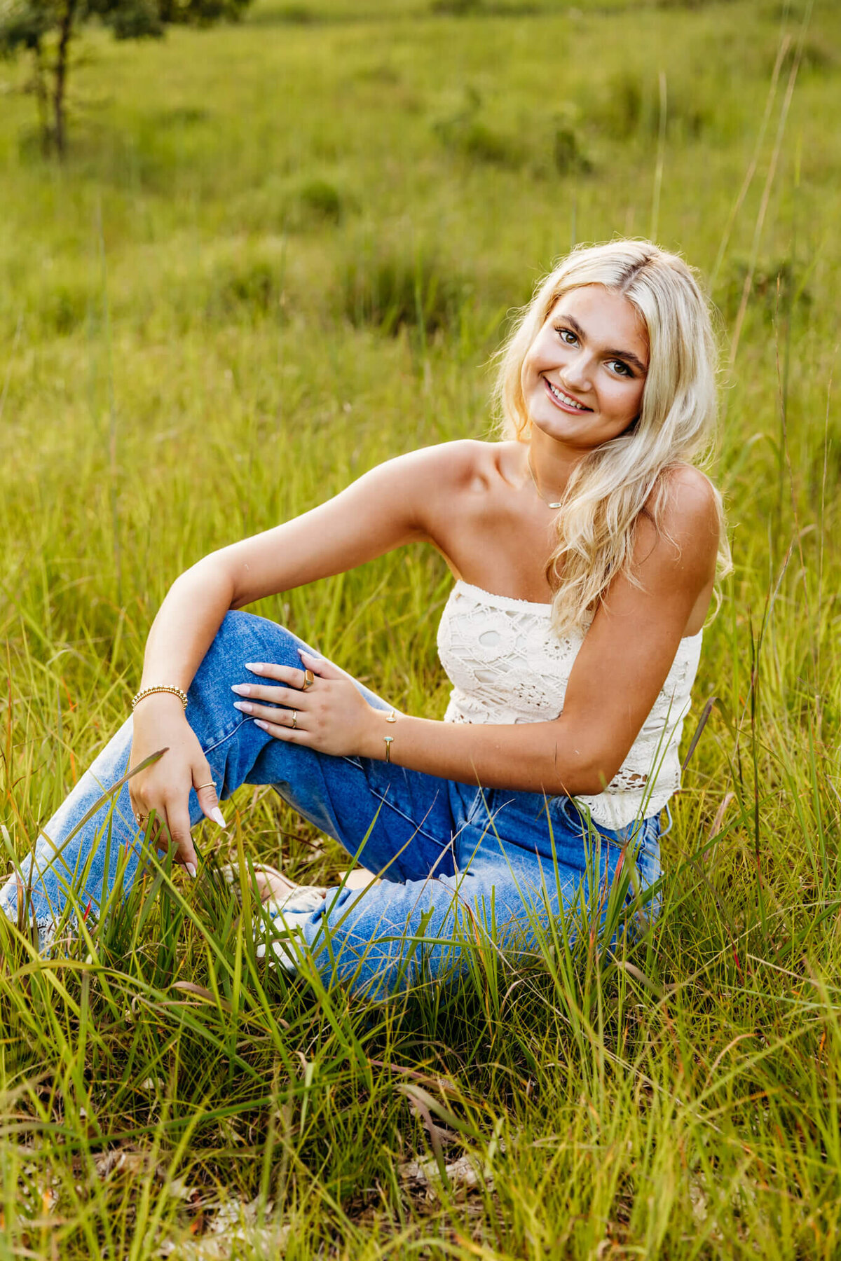 beautiful young woman sitting in a grassy field with one knee up and resting her elbow on it as she smiles for senior pictures.