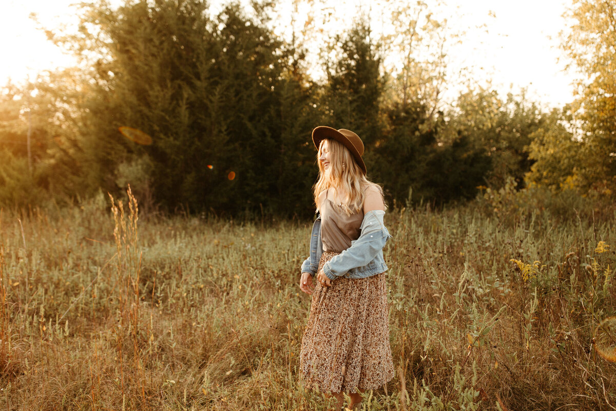 High school senior girl in long floral dress, wide brim hat, and denim jacket twirling in a field