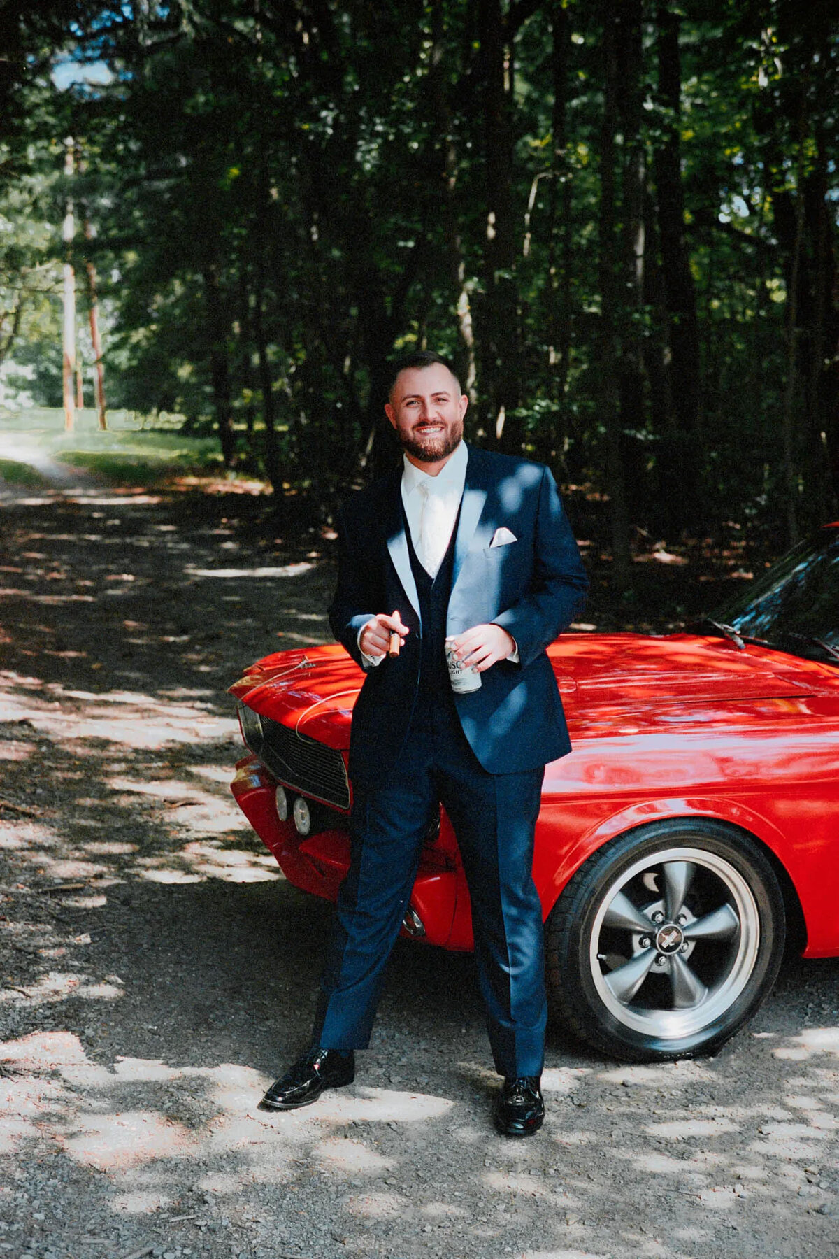 A man in a dark suit and white shirt stands smiling next to a red vintage car on a tree-lined, sun-dappled road. Captured by an NJ wedding photographer, he holds a drink confidently in the shade.