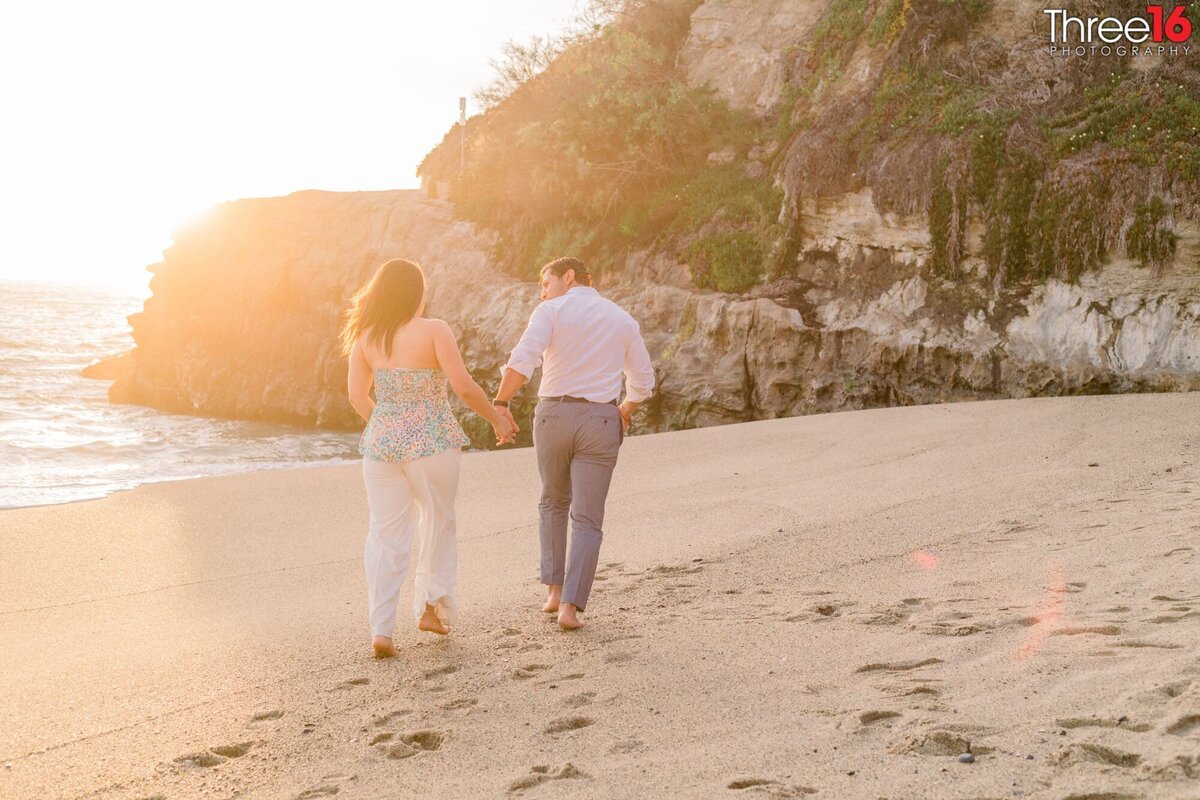 Newly engaged couple walk hand in hand along the beach