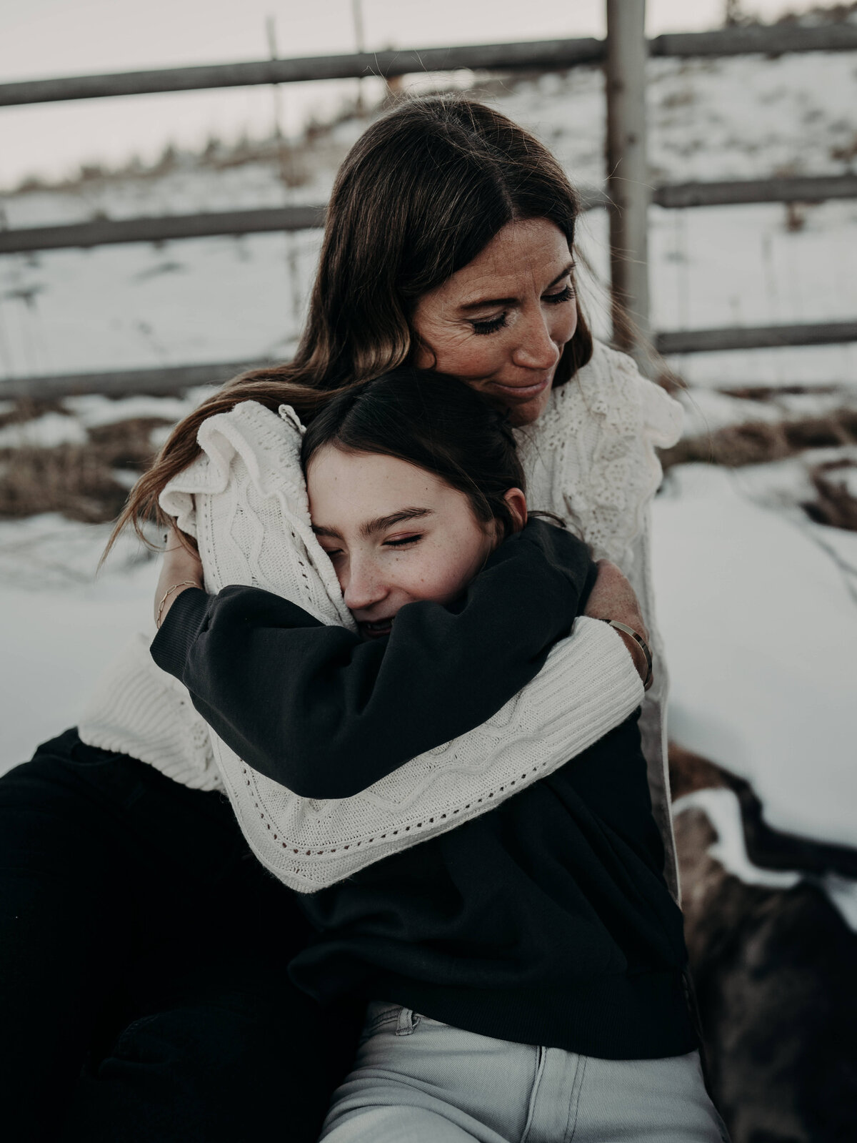 A mother prays over her teenage daughter as they hug sitting on a blanket with a snowy Colorado backdrop.