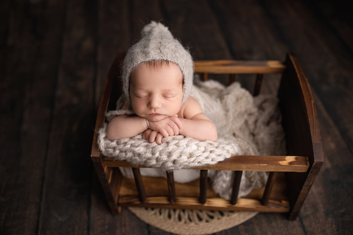Newborn boy posed in a wooden cradle wearing a tan bonnet.