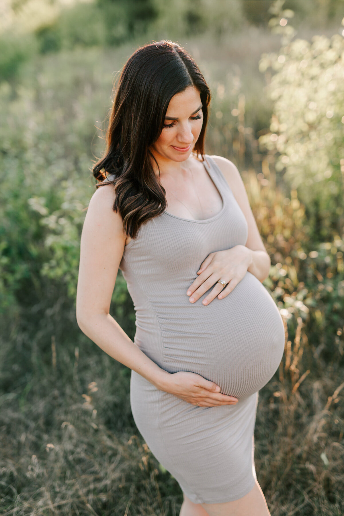 woman in taupe fitted dress looks down at her pregnant belly.