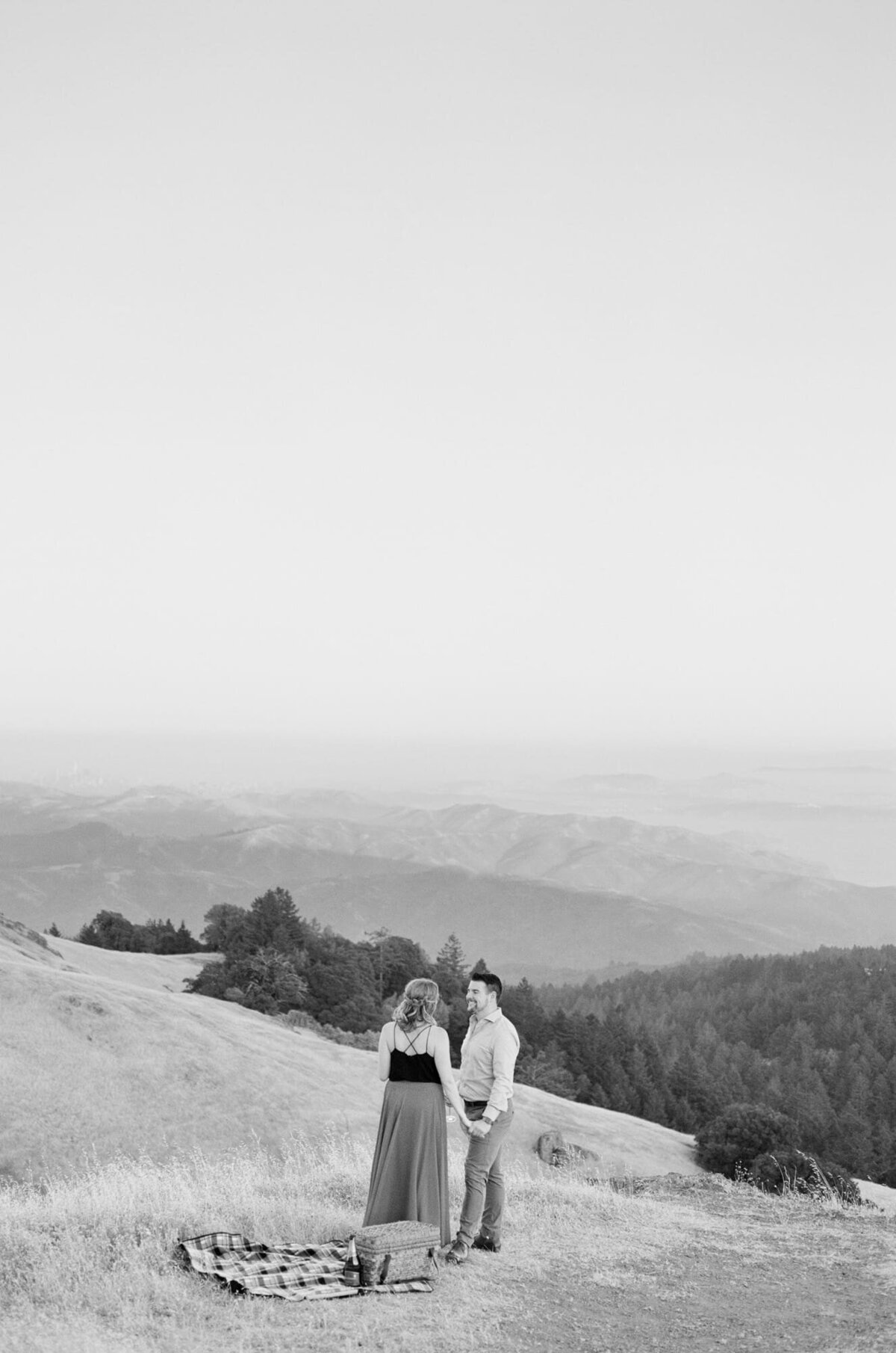 Adorable young couple enjoy a moment together with a picnic basket and picnic rug at a hilltop in front of hill ranges that seem to go on forever in  the background.