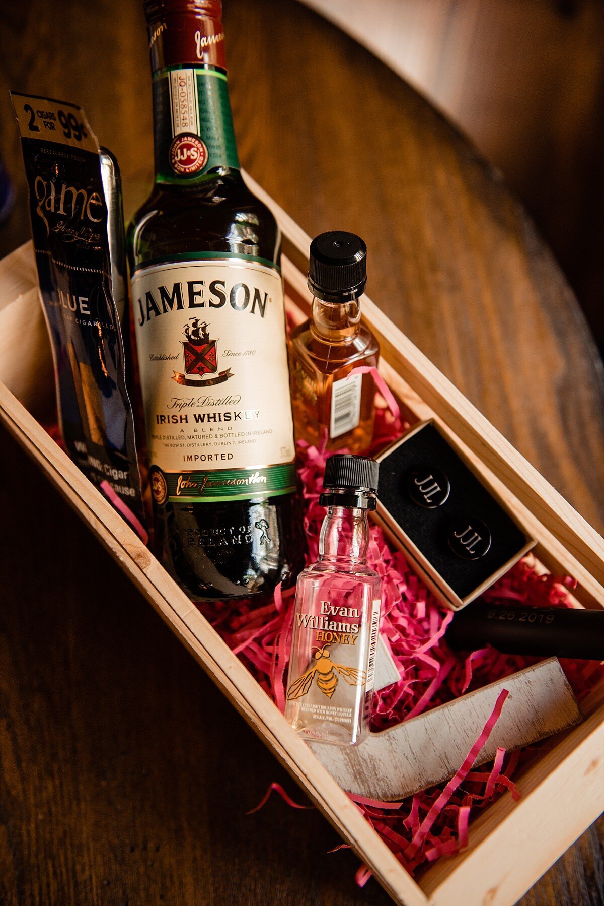 A gift basket for groomsmen with Irish Whisky, cufflinks and other items on a bed or red shredded paper in a wooden box.