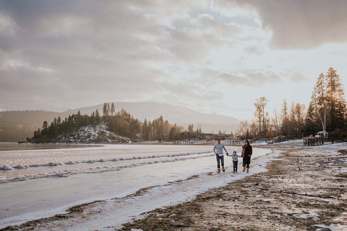 Family of three walk along the frozen shore of lake Pend Oreille while sun shines through clouds in the background.