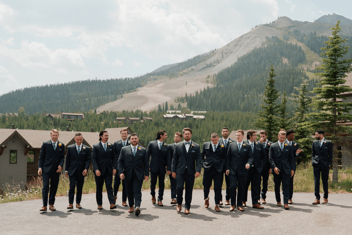 Groom and groomsmen walking together in navy suits at a summer wedding at Moonlight Basin, with forest and mountain views in the background.