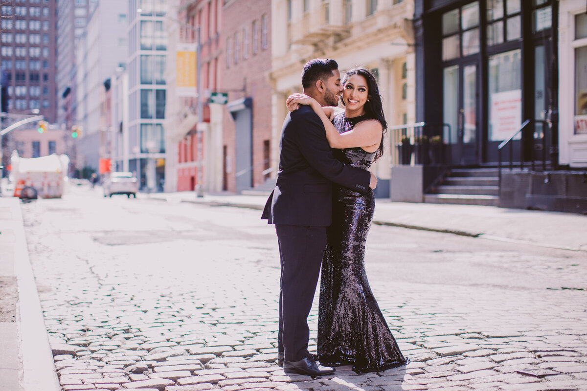 Groom kissing bride wearing black formal outfits on the cheek during their engagement shoot on a cobblestone street in soho new york
