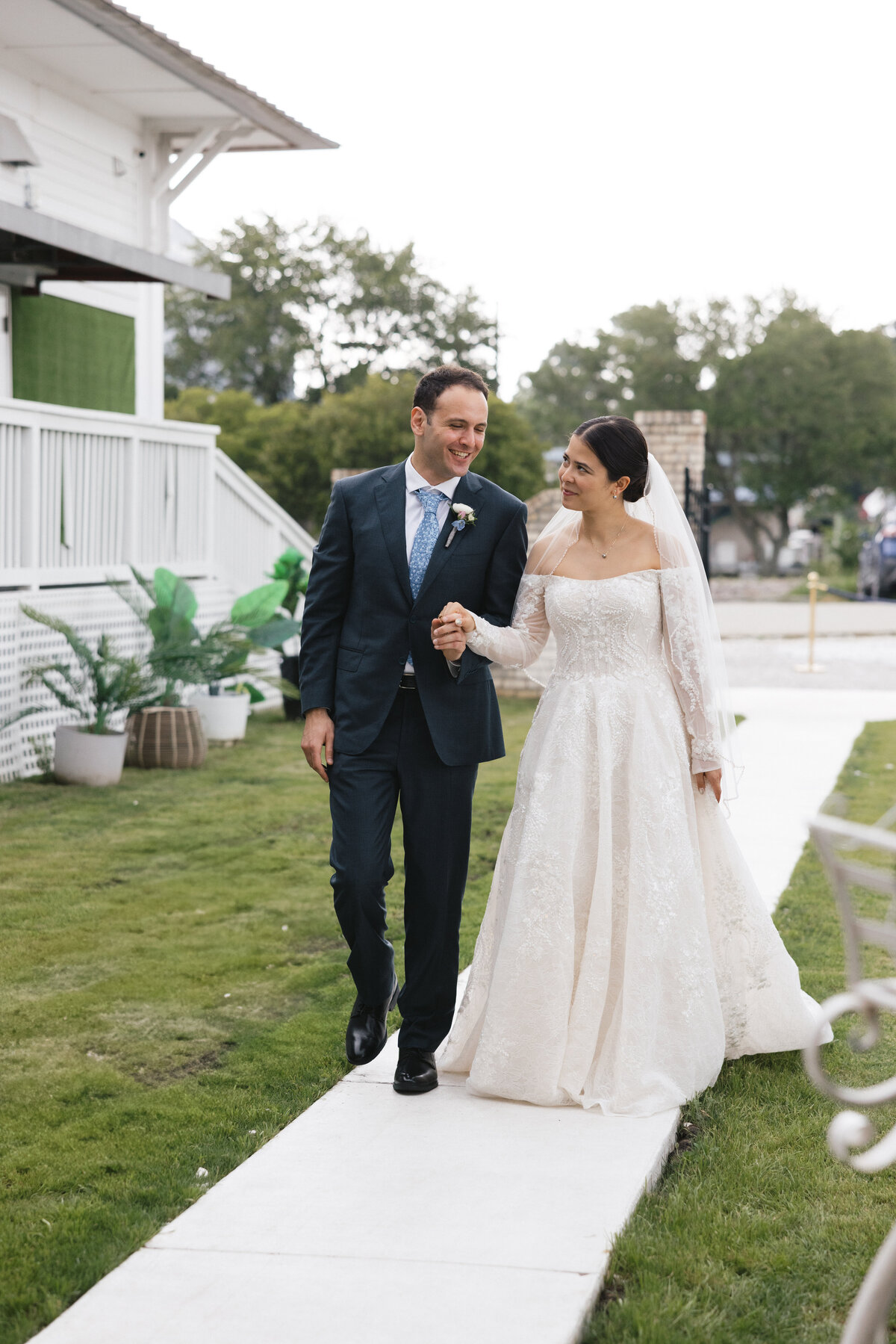 Bride and groom walking arm-in-arm along a garden path during their outdoor wedding, smiling at each other.
