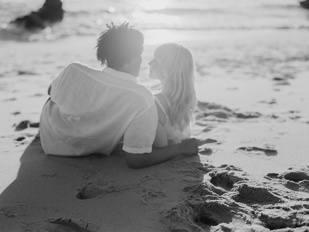 black and white image of couple snuggling at wind n sea beach in the sand laying down with backs towards the camera