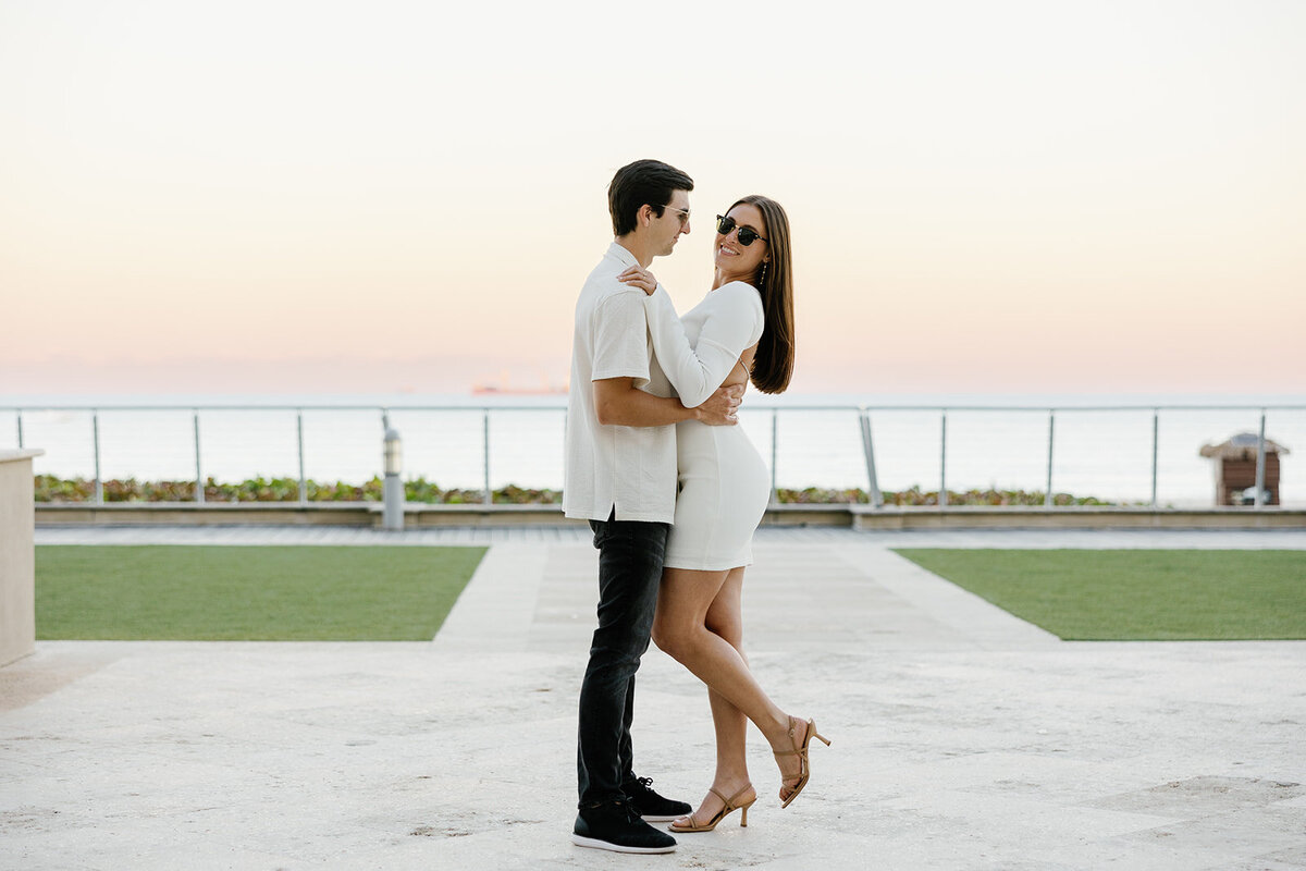 Graysen wraps his arms around Rebecca as they stand on the lush green lawn of Marriott Hotel Fort Lauderdale, savoring the joy of their engagement session with Claudia Amalia Photography.