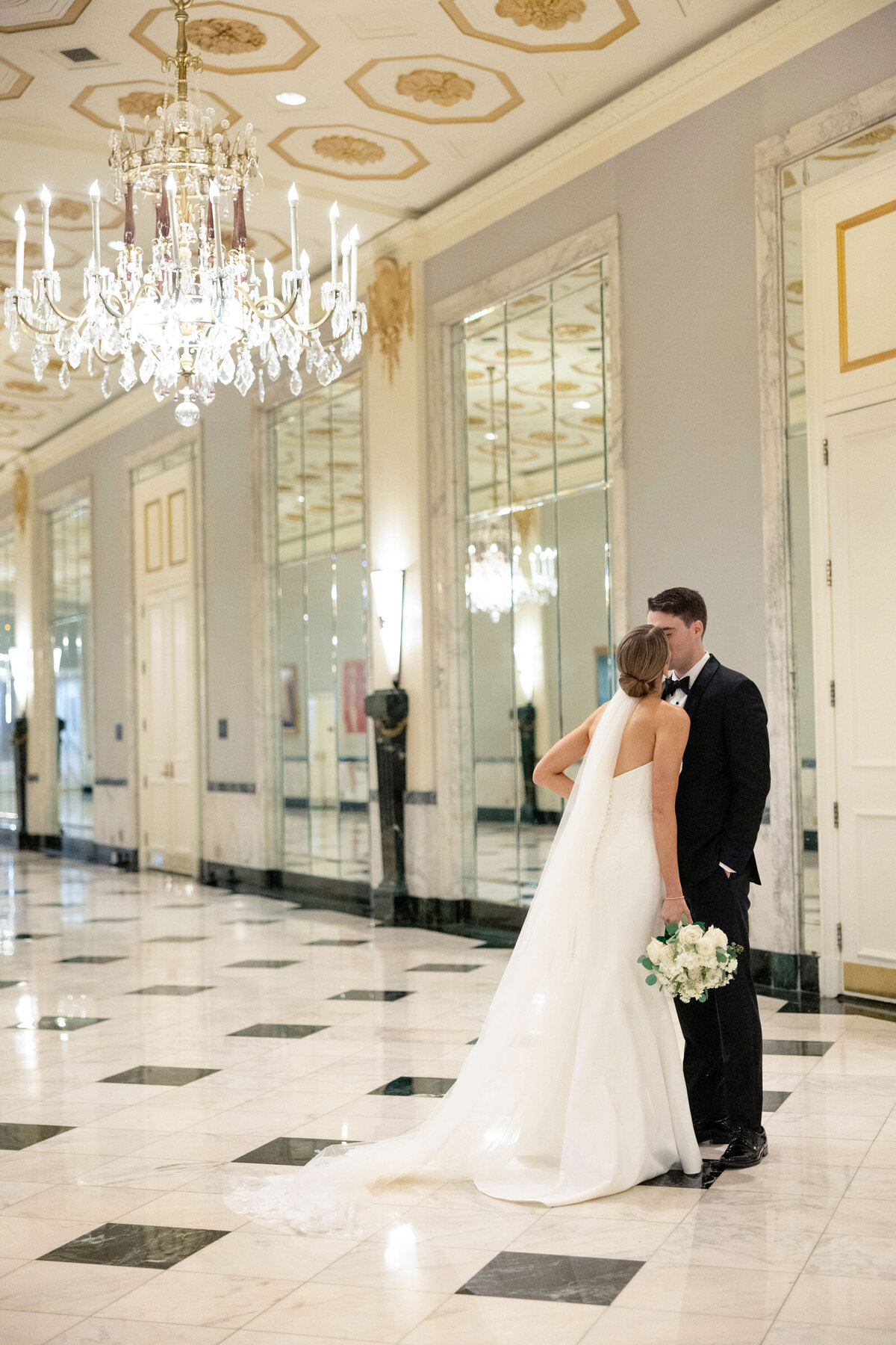 A bride and groom stand close in a luxurious hallway with a marble floor and ornate ceiling. The bride, in a flowing white gown, holds a bouquet as they kiss near large mirrors and a chandelier.