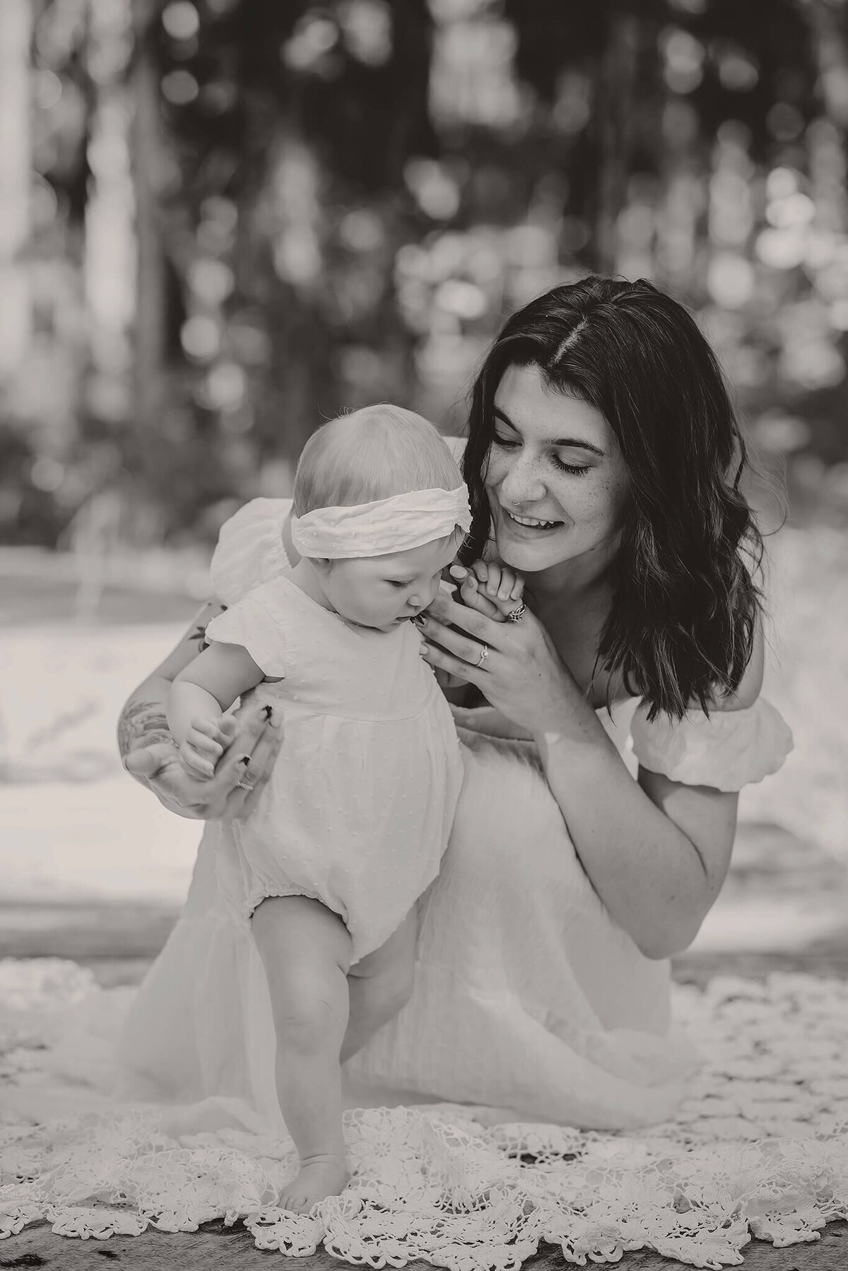 A mother and daughter share an embrace on a little wooden bridge. Mom is crouched down to hang onto baby girl who is standing. Mom looks lovingly at her. Black and white portrait by Brittany Danielle Photography
