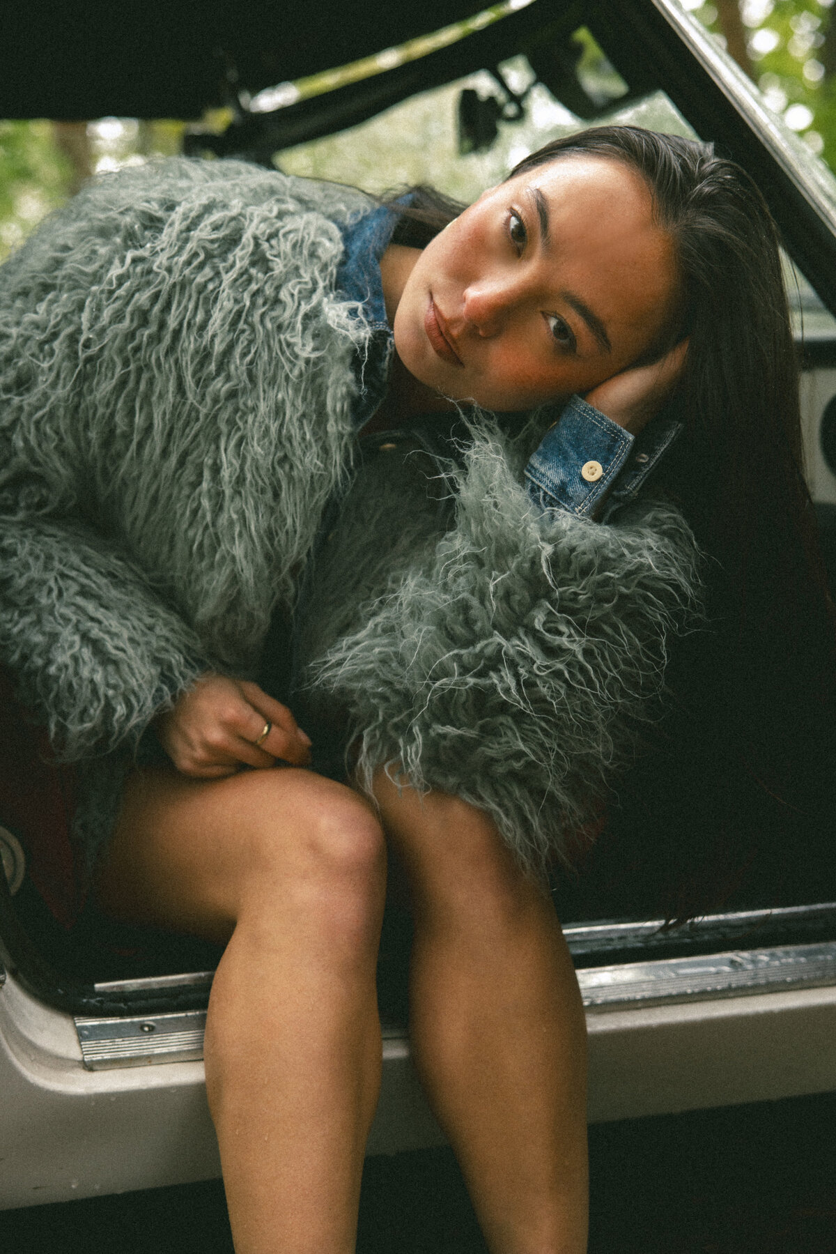 Sitting confidently inside the car, the model exudes a strong and stylish vibe, her fur coat and denim skirt perfectly complementing the vintage red leather seats. The use of lighting adds depth and mood to the composition. Photographed by Bo Eppink.