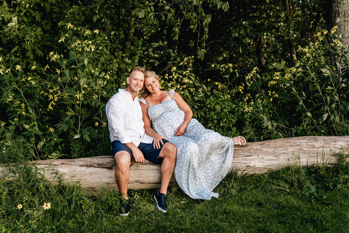 The couple sits on a fallen log in a forested area, surrounded by wildflowers and greenery.