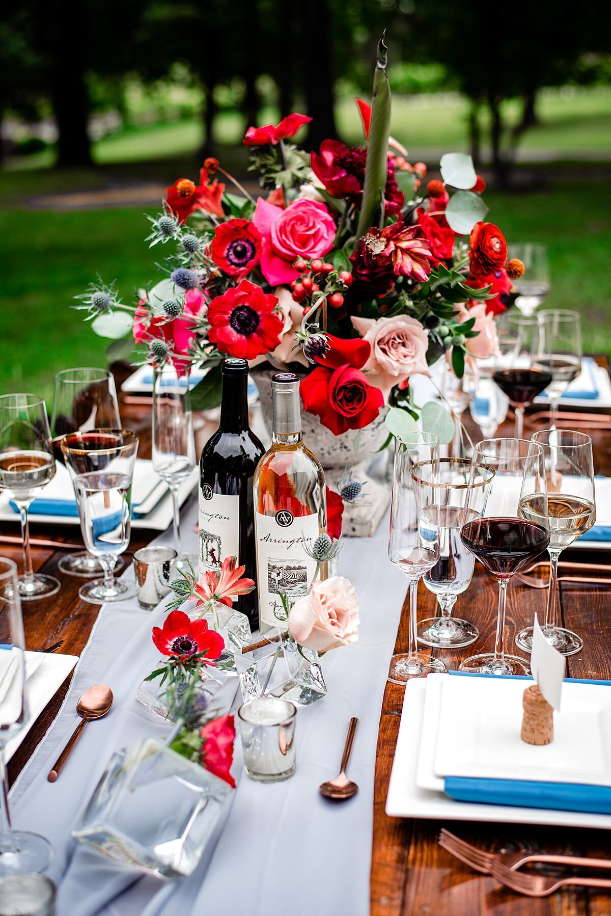 A farm table with a large red floral centerpiece featuring blush roses, red roses, red anthurium, red ranunculus, red anemones, blush and red protea, blue thistle, hypericum berries, red cascading dianthus and assorted greenery. The dark wood farm table is also set with a dove gray sheer organza table runner and cube shaped glass bud vases with red and blush flowers. The matte copper flatware is set on either side of the square white dinner plates that are topped with salad plates wrapped in teal linen napkins, and white square dessert plates. Wine cork seating card stands are on each plate with a white seating card with teal calligraphy. The table is also set with red and white tulip shaped long stemmed wine glasses and footed crystal water goblets with a gold rim. Bottles of Arrington Vineyards Stag's White, and Red Fox Red wines are set on the table.