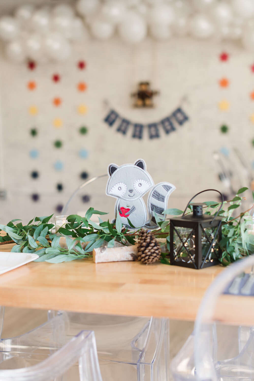 Woodland-themed table centerpiece for a baby boy shower featuring wood logs, lush greenery, a lantern, and a fabric squirrel.