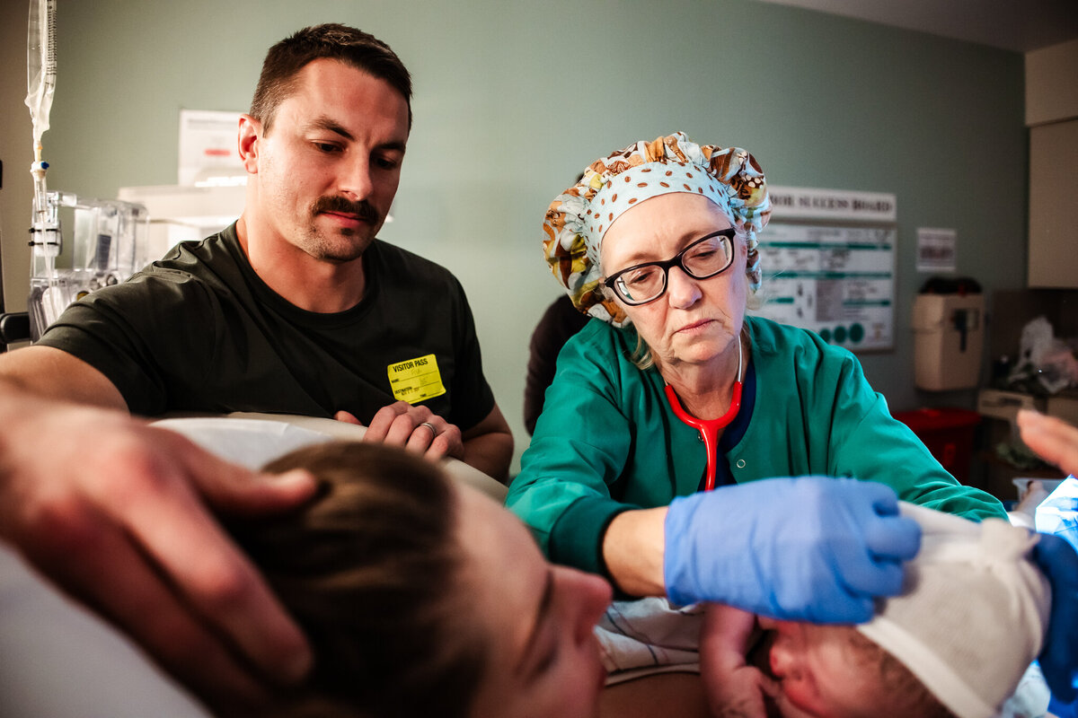 A father and nurse look down at the newly born baby as he cuddles into his mother.  