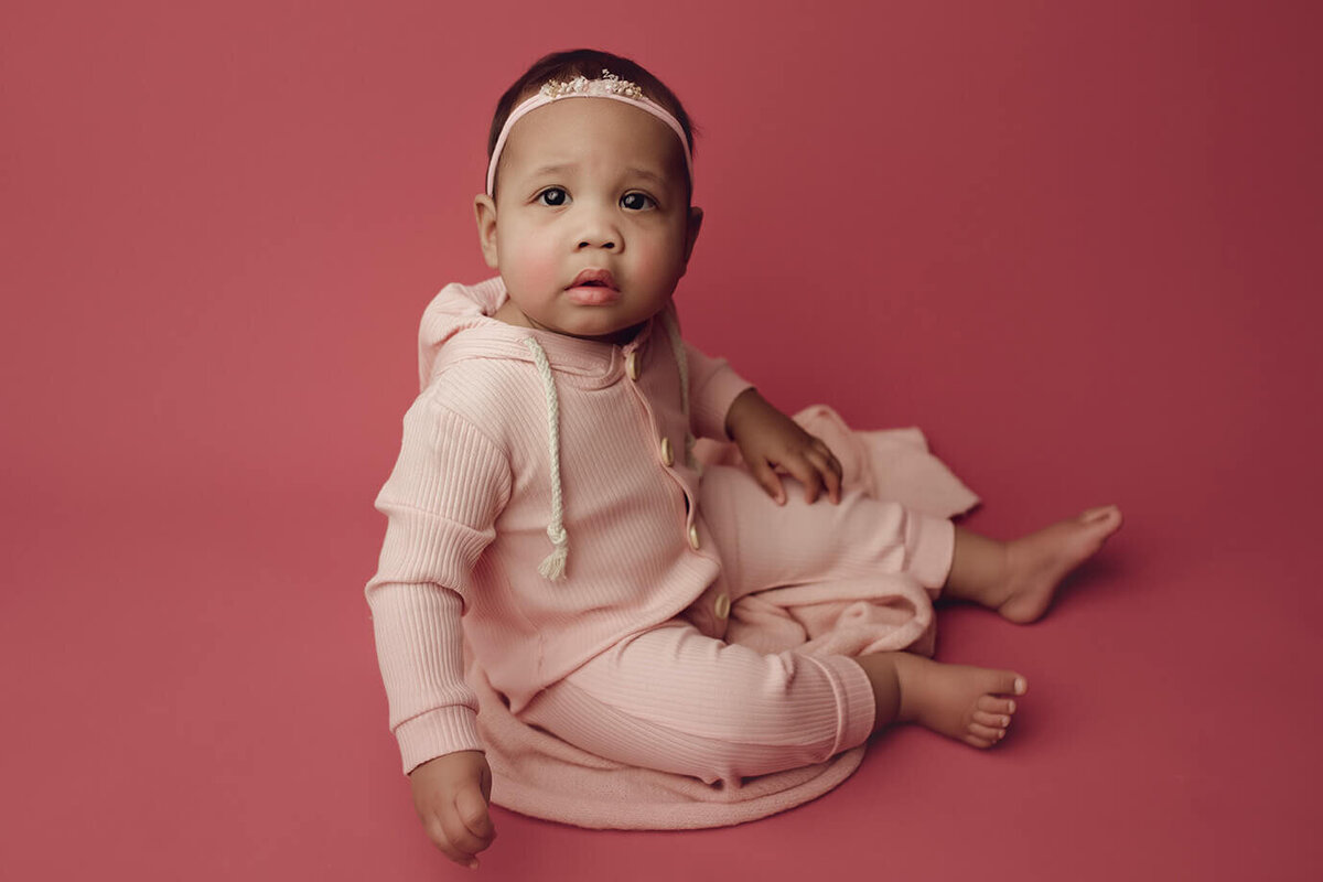 a one year old girl sitting on a pink wrap in a pink outfit on a pink backdrop