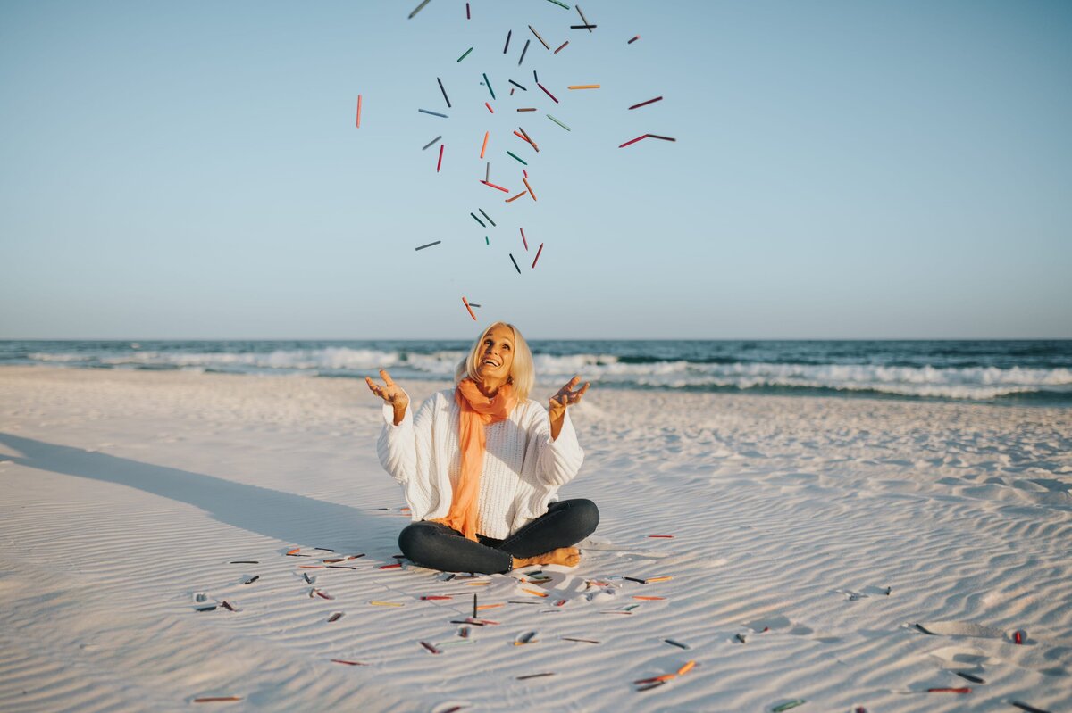 Older woman with white hair wearing a white long-sleeve shirt, jeans and an orange scarf sits cross-legged on the sand at the beach and throws colorful crayons in the air.
