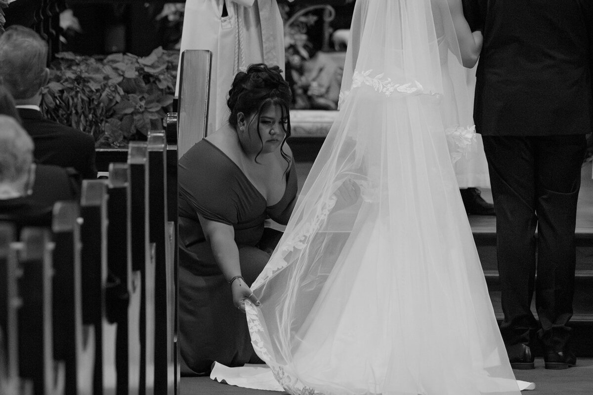 Editorial capture of a maid of honor adjusting and straightening designer gown and veil for bride at the front of San Antonio church wedding