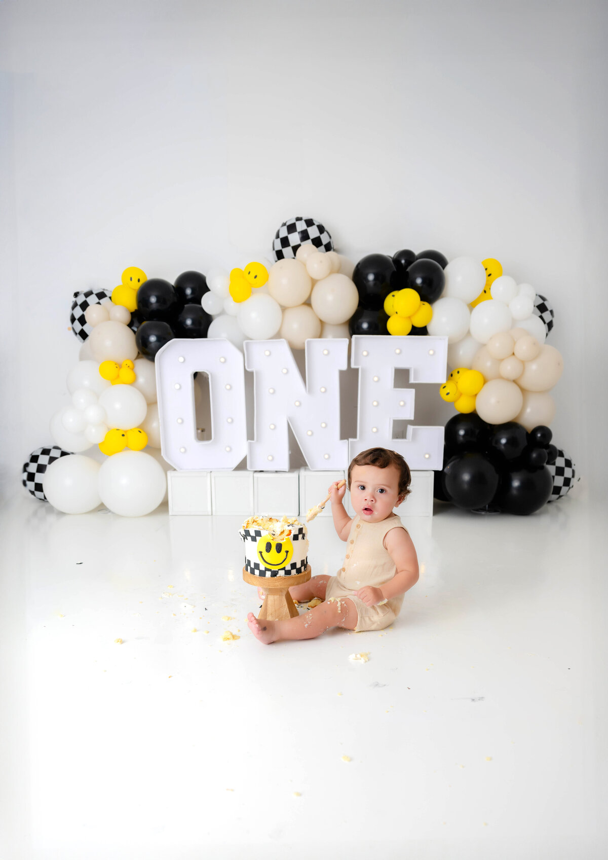 One-year-old boy with cake between his legs, surrounded by "ONE" letters and colorful balloons.