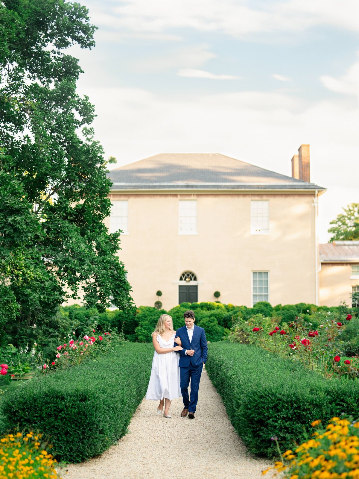 A couple walks arm in arm down a gravel path in a lush garden with blooming flowers on either side. They are in front of a large beige house with a gray roof, surrounded by greenery under a partly cloudy sky.