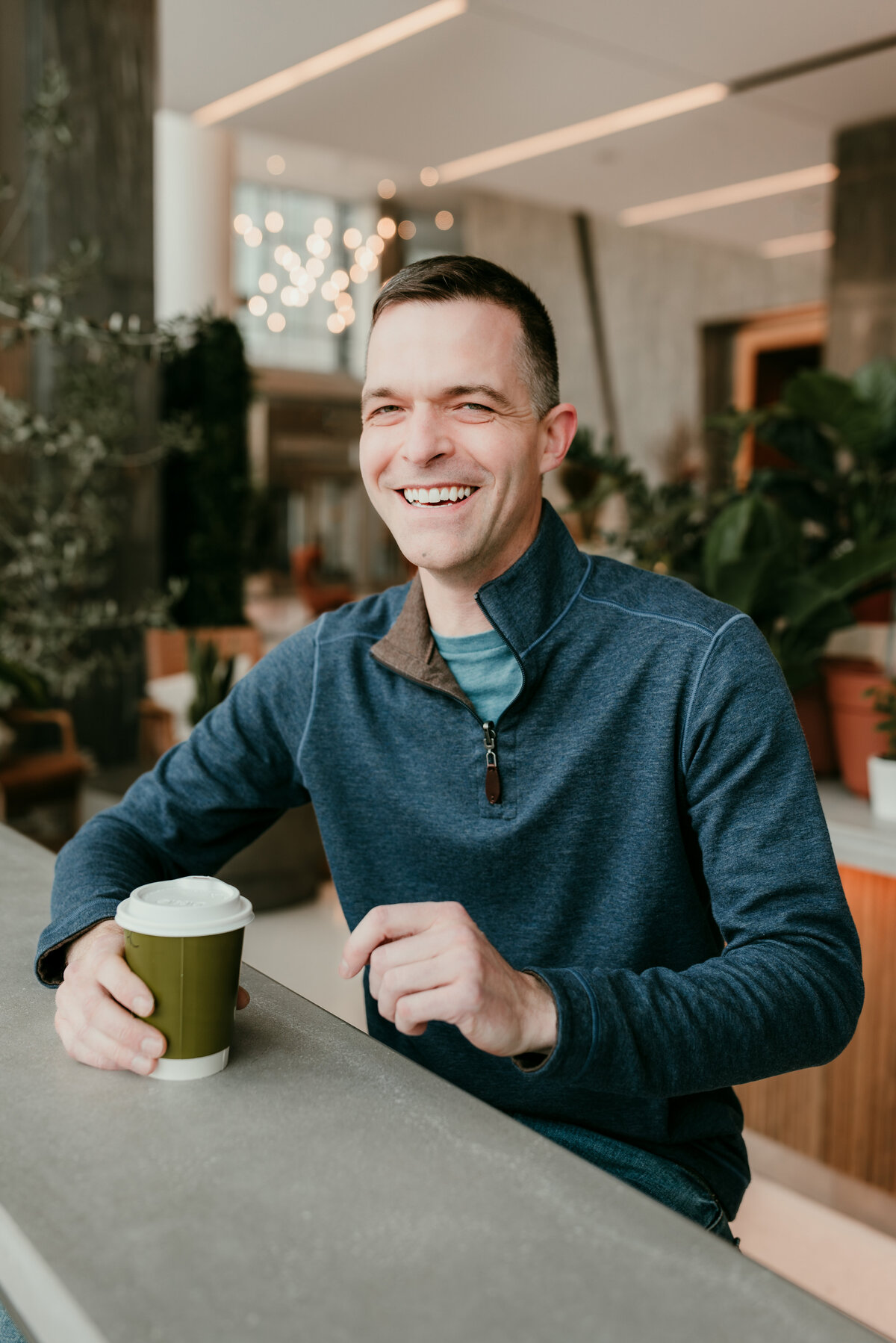 Man smiling warmly while holding a coffee cup in a modern coffee shop, radiating confidence and approachability for an online dating profile.