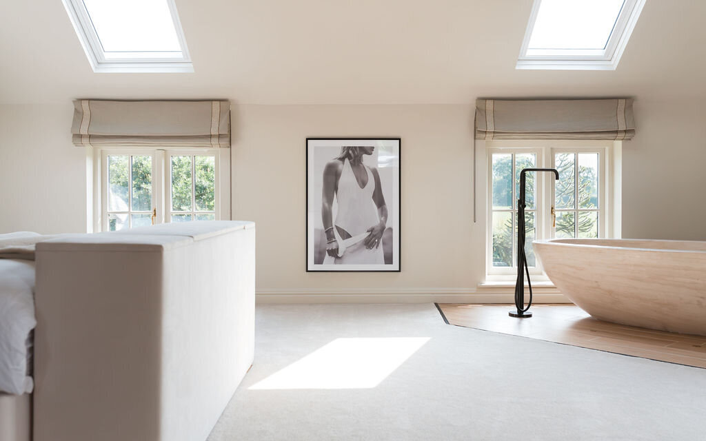 A serene, minimalistic bedroom features a large bathtub with modern fixtures, and twin skylights. A framed monochromatic photograph hangs between two sets of windows, adding a contemporary touch to the airy space.