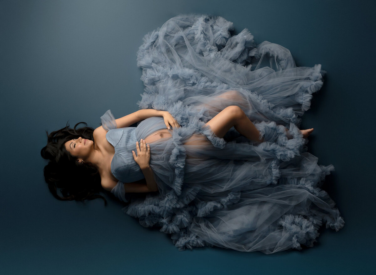 Pregnant woman lying on floor in flowing blue ruffled maternity gown in studio portrait