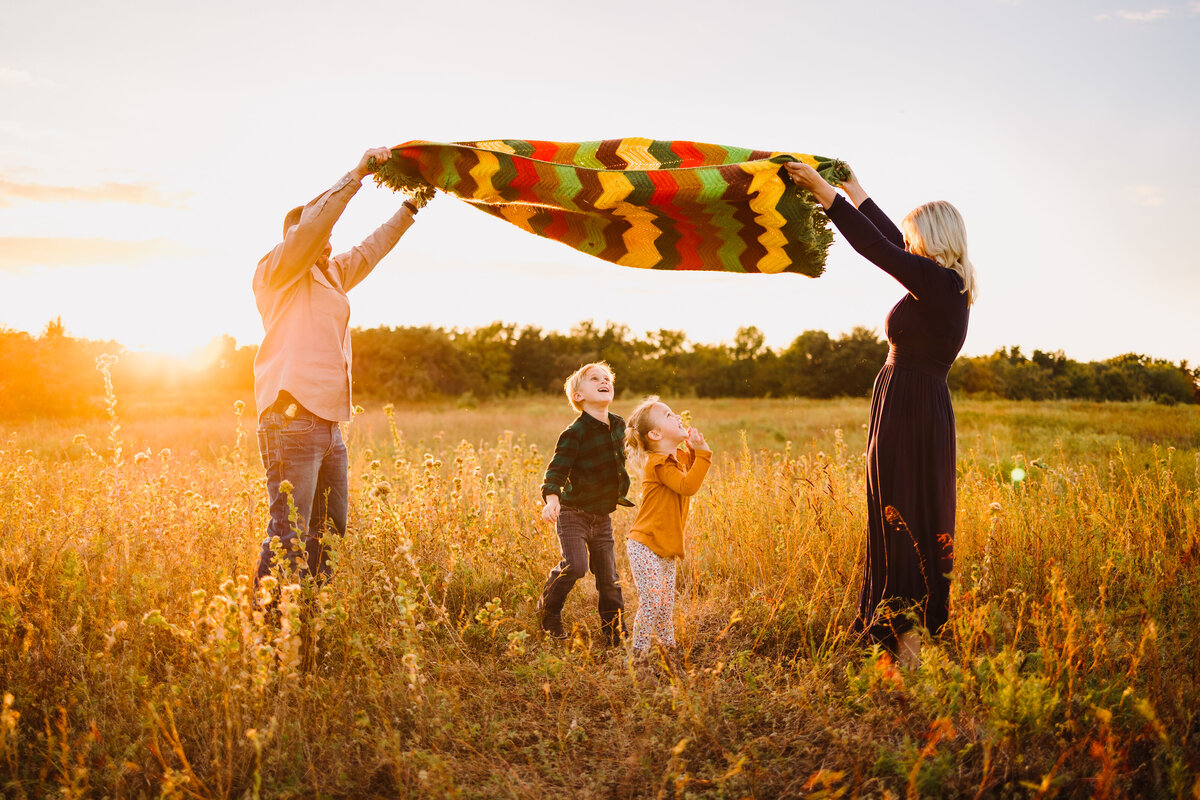 Family photo in a garden, the woman in a long dress is holding with the man in a shirt a green, orange, and yellow blanket above her head. The children are looking up and smiling because they have fun