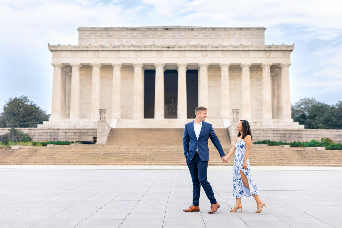 A couple holding hands and smiling at each other in front of the Lincoln Memorial. The man is wearing a blue suit, and the woman is in a blue and white floral dress. The sky is slightly overcast.